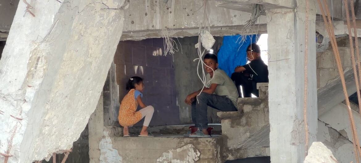 A young girl crouches and a boy sits on broken concrete steps inside a war-damaged building in Gaza City, with an adult seated nearby amid rubble and exposed rebar.