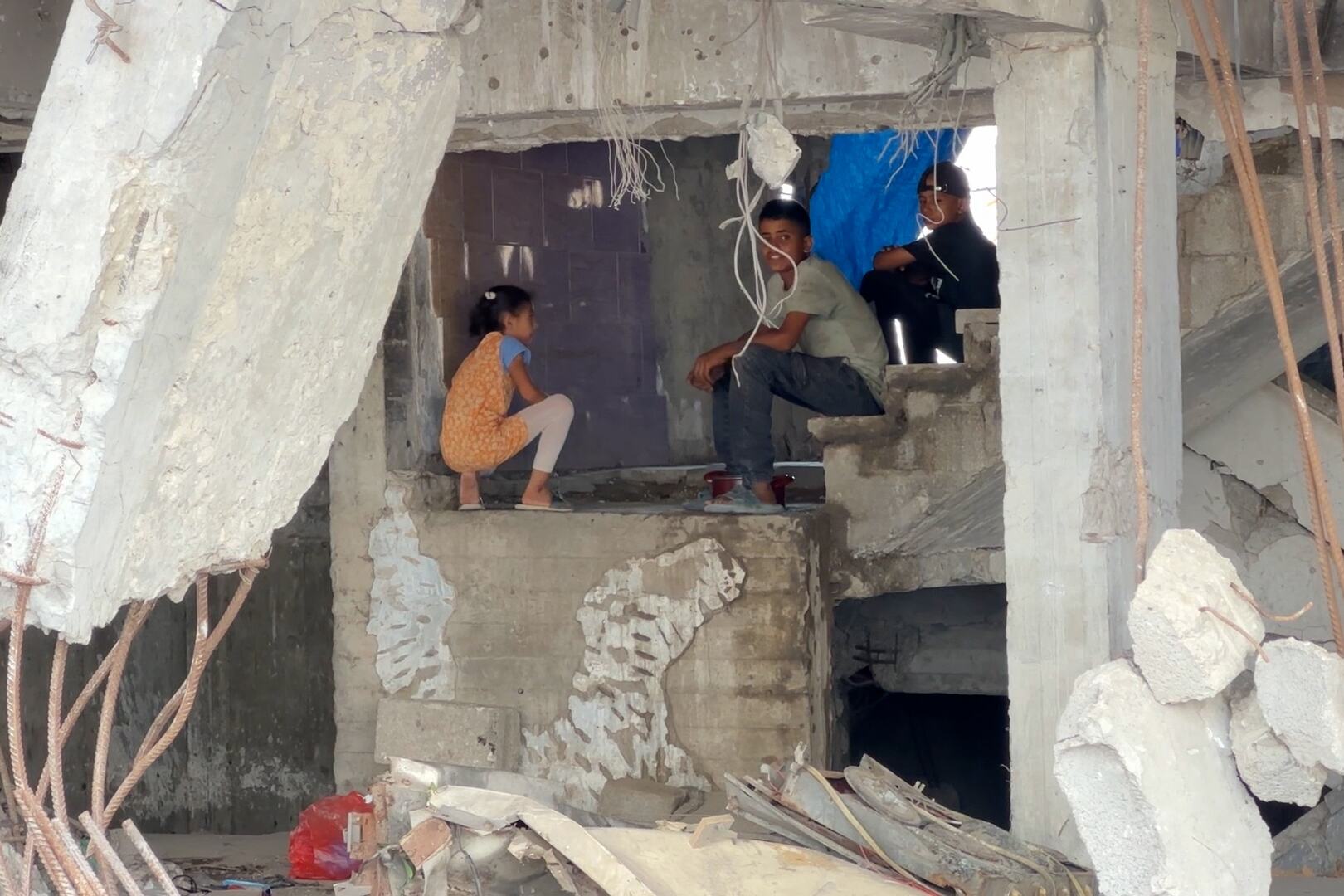 فتاة صغيرة تجلس القرفصاء وفتى يجلس على درجات خرسانية مكسورة داخل مبنى متضرر من الحرب في مدينة غزة، بينما يجلس شخص بالغ بالقرب منها وسط الأنقاض وقضبان التسليح المكشوفة.