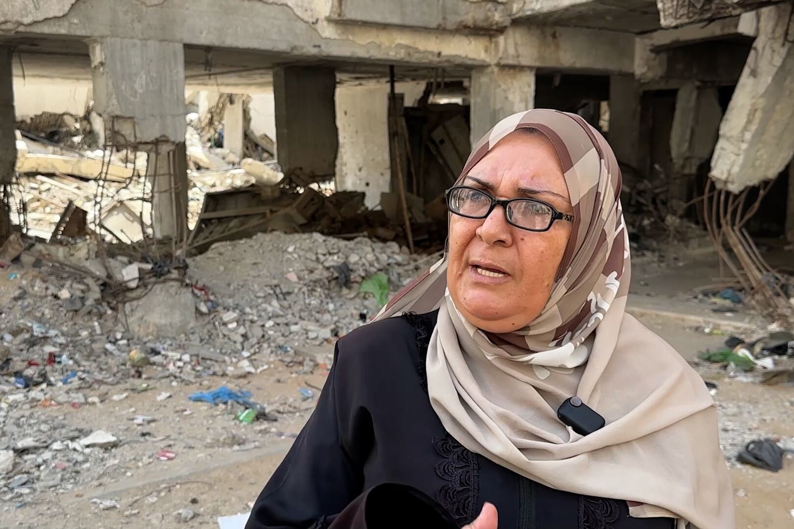 تقف أم محمد أمام مبنى خرساني متضرر تظهر فيه قضبان التسليح والأنقاض، وتتحدث عن منزلها المعرض لخطر الانهيار في حي الكرامة بمدينة غزة.