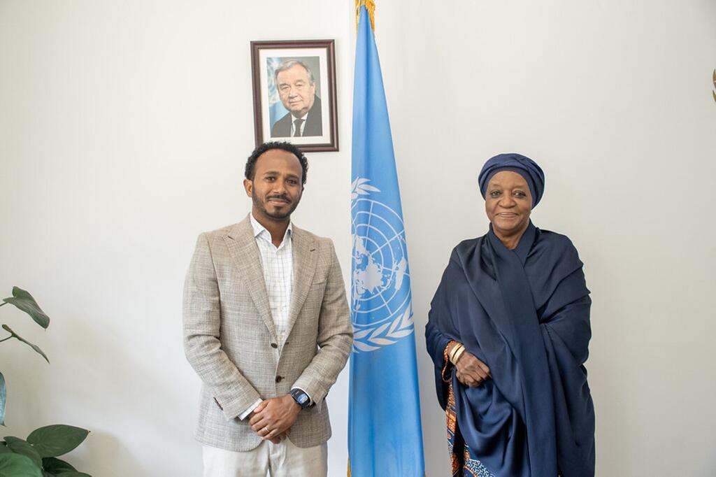 Un homme en costume beige et une femme en tenue traditionnelle bleu marine se tiennent à côté d'un drapeau des Nations Unies dans un bureau où un portrait encadré est accroché au mur.