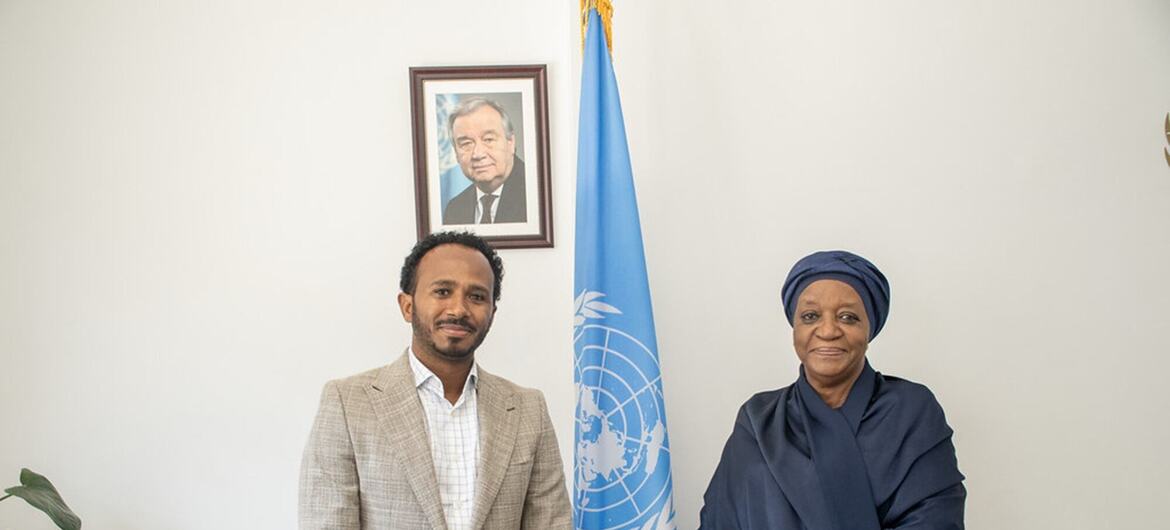 A man in a beige suit and a woman in navy traditional attire stand beside a United Nations flag in an office with a framed portrait on the wall.