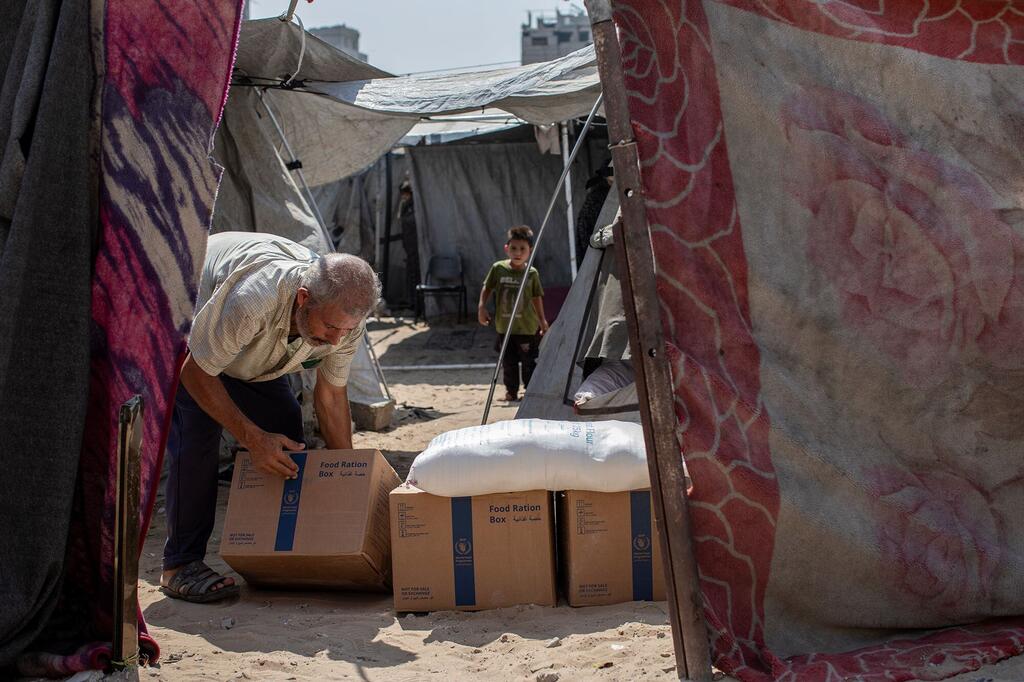 Un homme âgé du Moyen-Orient se penche sur des boîtes de rations alimentaires du Programme alimentaire mondial et un sac blanc dans un camp de tentes sablonneux, tandis qu'un jeune garçon se tient debout parmi des abris en bâche.
