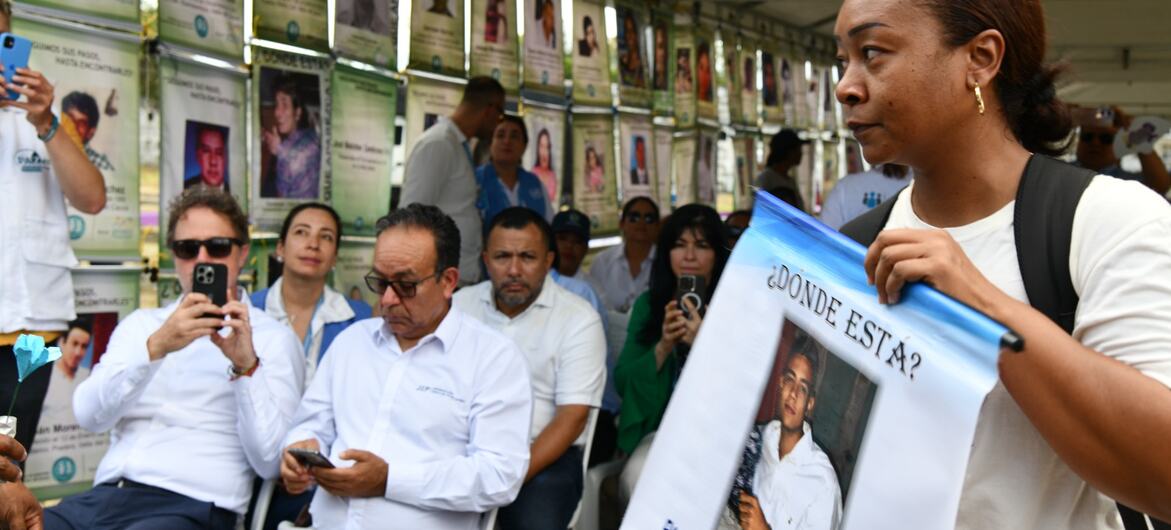 Una mujer sostiene un cartel con un retrato que dice "¿Dónde está?" durante una reunión al aire libre bajo una carpa, con filas de retratos de personas desaparecidas exhibidas en una pared detrás de los asistentes sentados.