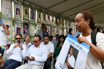 Une femme tient une pancarte avec un portrait sur laquelle on peut lire « ¿Dónde está? » lors d'un rassemblement en plein air sous une tente, avec des rangées de portraits de personnes disparues affichées sur un mur derrière les participants assis.