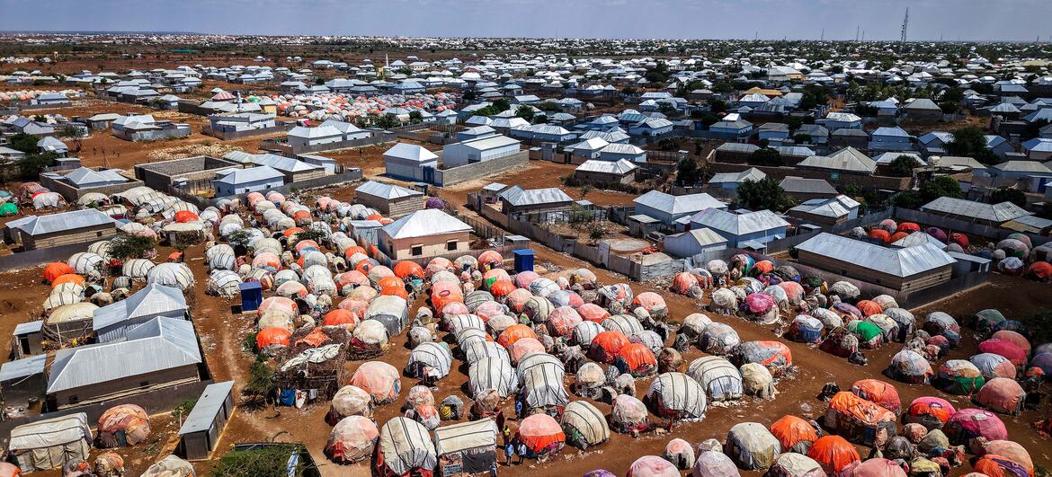Aerial view of Baidoa's displacement camps in Somalia showing dense rows of tarpaulin tents and metal-roofed shelters across dry land under a partly cloudy sky. Aerial view of Baidoa's displacement camps in Somalia showing dense rows of tarpaulin tents and metal-roofed shelters across dry land under a partly cloudy sky.