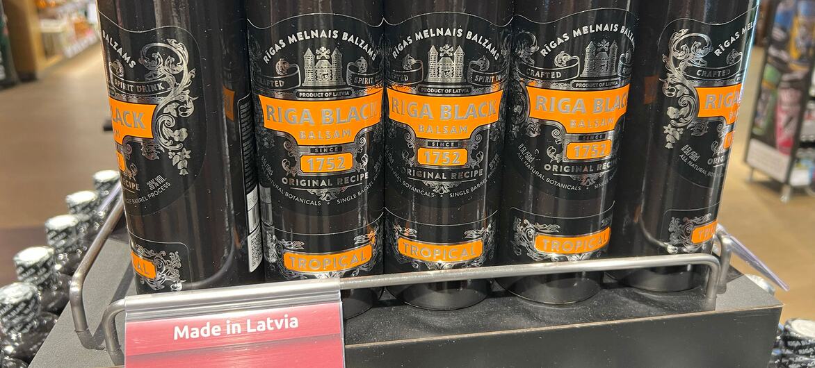 Rows of dark Riga Black Balsam bottles are arranged on a supermarket shelf, with a red and white Made in Latvia sign displayed in front.