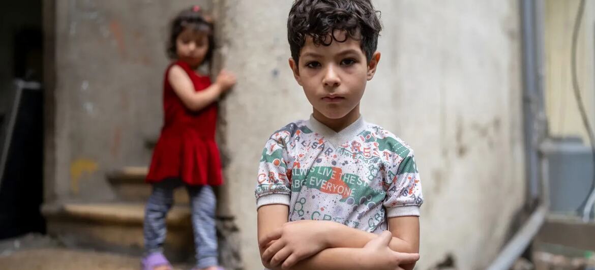 Um menino de braços cruzados está em pé em frente a uma parede de concreto desgastada, enquanto uma menina de vestido vermelho se encosta na parede atrás dele.
