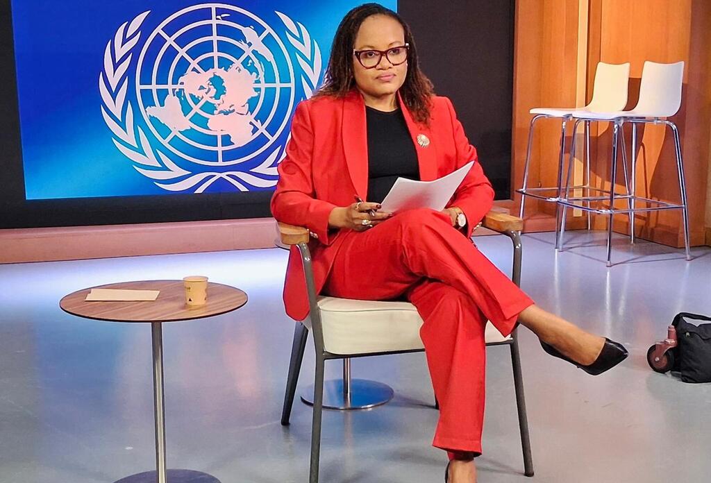 Mwanamke aliyevaa suti nyekundu ameketi kwenye kiti cha studio akiwa ameshika karatasi, huku nembo UMOJA WA MATAIFA ikiwa imeonyeshwa kwenye skrini ya bluu nyuma yake.