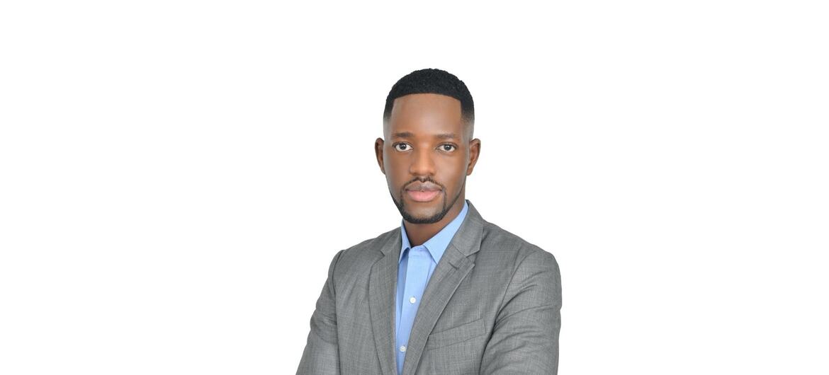 Paul kutoka Tanzania amevalia suti ya kijivu na shati la bluu nyepesi huku mikono yake ikiwa imevuka dhidi ya mandhari nyeupe isiyo na dosari.