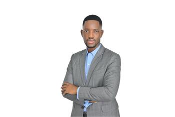Paul kutoka Tanzania amevalia suti ya kijivu na shati la bluu nyepesi huku mikono yake ikiwa imevuka dhidi ya mandhari nyeupe isiyo na dosari.