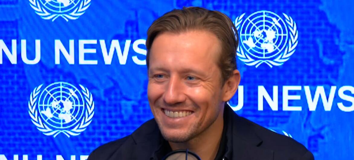 Lucas Leiva sorri enquanto fala ao microfone em frente a um painel azul com o logotipo das Nações Unidas e emblemas repetidos da organização.