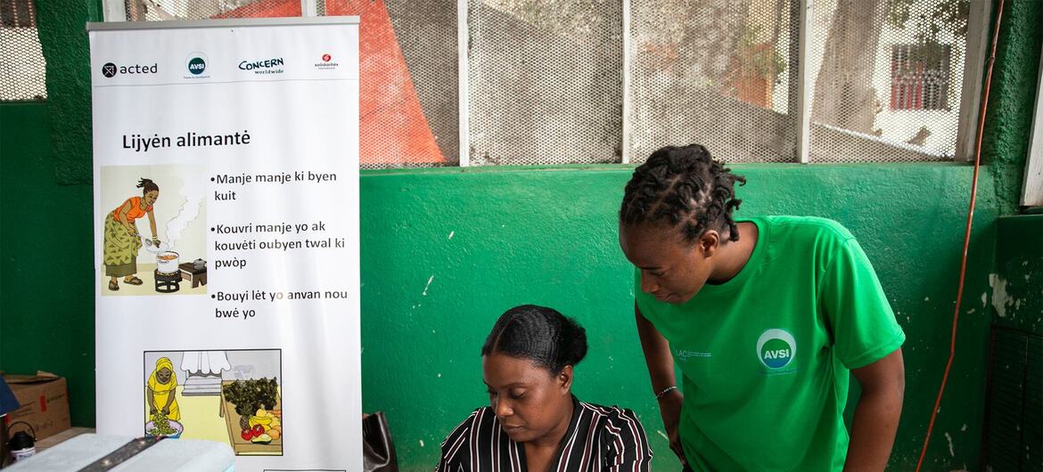 Une femme noire en chemisier rayé remplit des formulaires de santé à une table, assistée d'un jeune bénévole noir de l'AVSI en chemise verte ; une banderole haïtienne sur la sécurité alimentaire se dresse derrière eux.