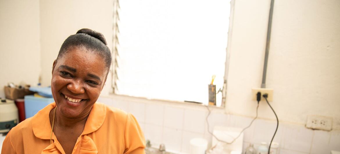 Une femme noire souriante, vêtue d'un chemisier orange et d'un gant jetable, tient un tube à essai au comptoir d'un laboratoire de clinique.