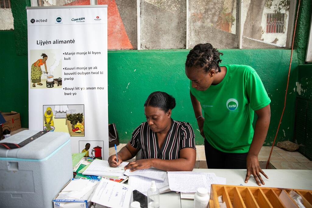 Une femme noire en chemisier rayé remplit des formulaires de santé à une table, assistée d'un jeune bénévole noir de l'AVSI en chemise verte ; une banderole haïtienne sur la sécurité alimentaire se dresse derrière eux.