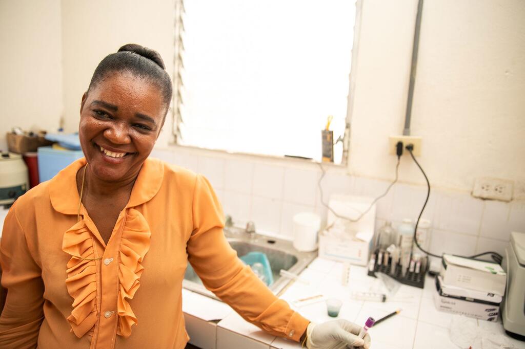 Une femme noire souriante, vêtue d'un chemisier orange et d'un gant jetable, tient un tube à essai au comptoir d'un laboratoire de clinique.