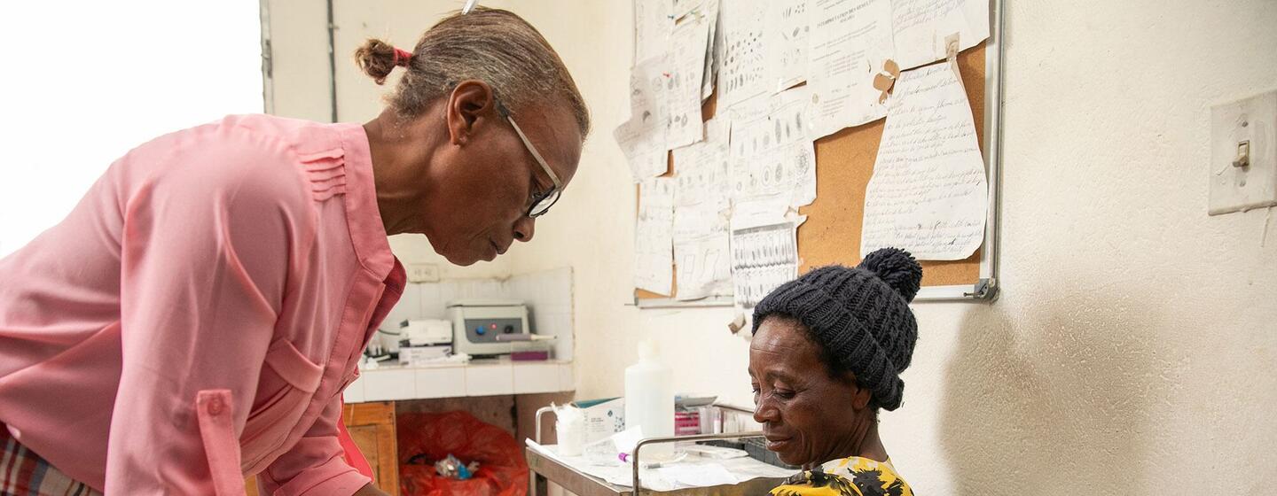 Una trabajadora sanitaria negra, vestida con una blusa rosa y guantes, prepara una vía intravenosa para una paciente negra sentada, que lleva un vestido amarillo con estampado floral y un gorro negro, dentro de una pequeña sala de consulta con material médico y avisos en papel.