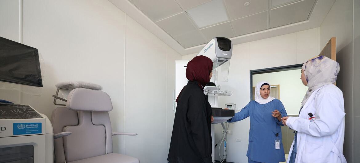 Three women in a hospital room with a mammography machine; one woman in black abaya and red hijab stands near the device while two female healthcare workers in blue scrubs and white lab coat explain its use.