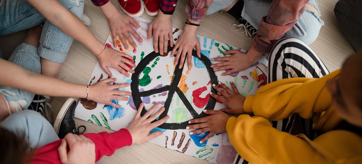 Vista de cima para baixo de várias crianças em idade escolar pressionando as mãos sobre um grande símbolo da paz de papel pintado com impressões de mãos multicoloridas no chão de uma sala de aula.