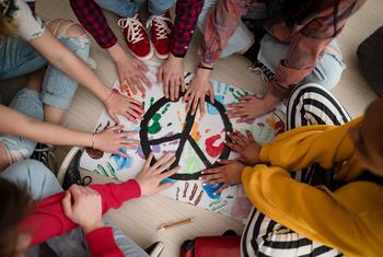 Vue plongeante de plusieurs écoliers appuyant leurs mains sur un grand symbole de paix en papier, peint d'empreintes de mains multicolores, sur le sol d'une salle de classe.
