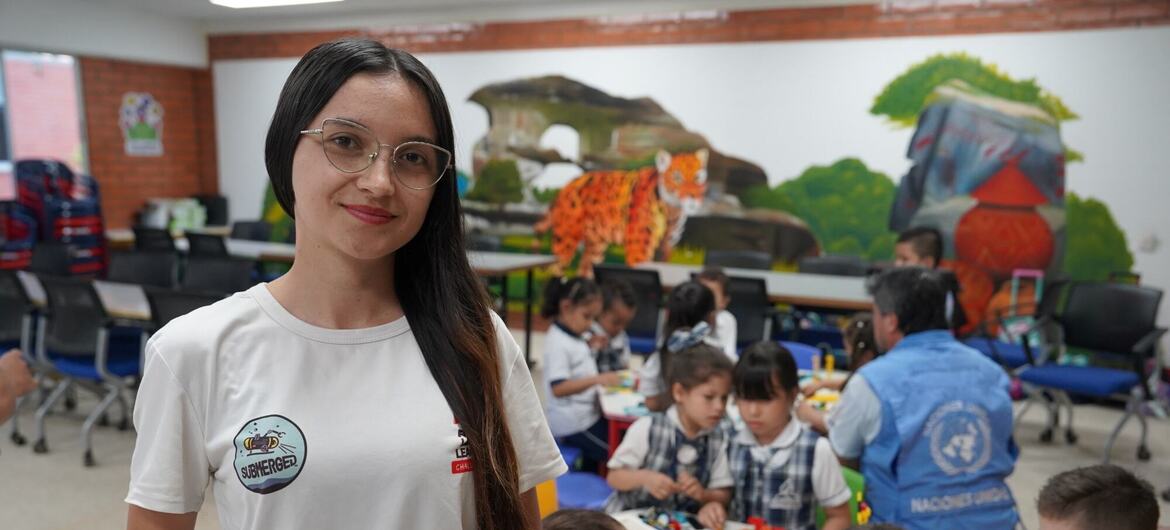 Una joven con gafas y una camiseta blanca con un logotipo aparece de pie sonriendo en un aula, mientras los niños realizan manualidades en las mesas situadas detrás de ella, bajo un colorido mural de un jaguar.