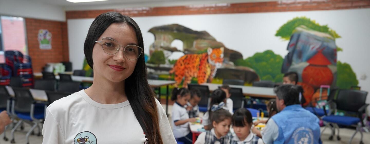 Una joven con gafas y una camiseta blanca con un logotipo aparece de pie sonriendo en un aula, mientras los niños realizan manualidades en las mesas situadas detrás de ella, bajo un colorido mural de un jaguar.