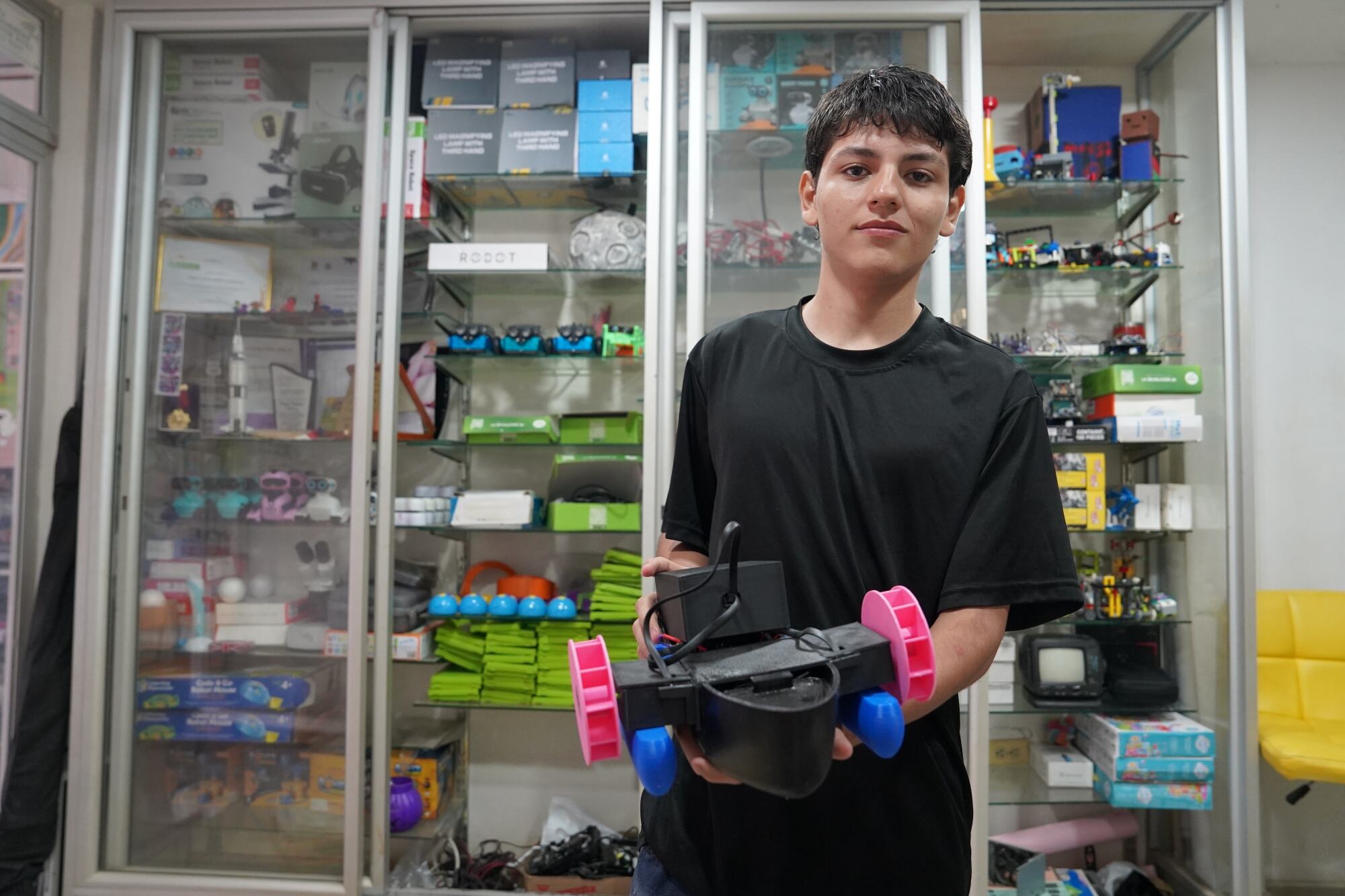 Un joven estudiante latino, vestido con una camiseta negra, sostiene un pequeño robot con ruedas rosas y azules frente a vitrinas llenas de kits educativos.