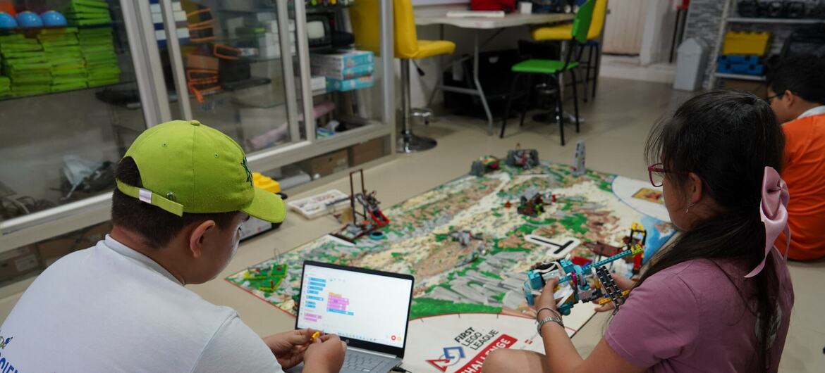 Dos jóvenes estudiantes, un niño con una gorra verde y una niña con un lazo rosa, están sentados en el suelo de un aula de robótica usando un ordenador portátil y montando un robot LEGO junto a una alfombrilla de juego de FIRST Challenge.
