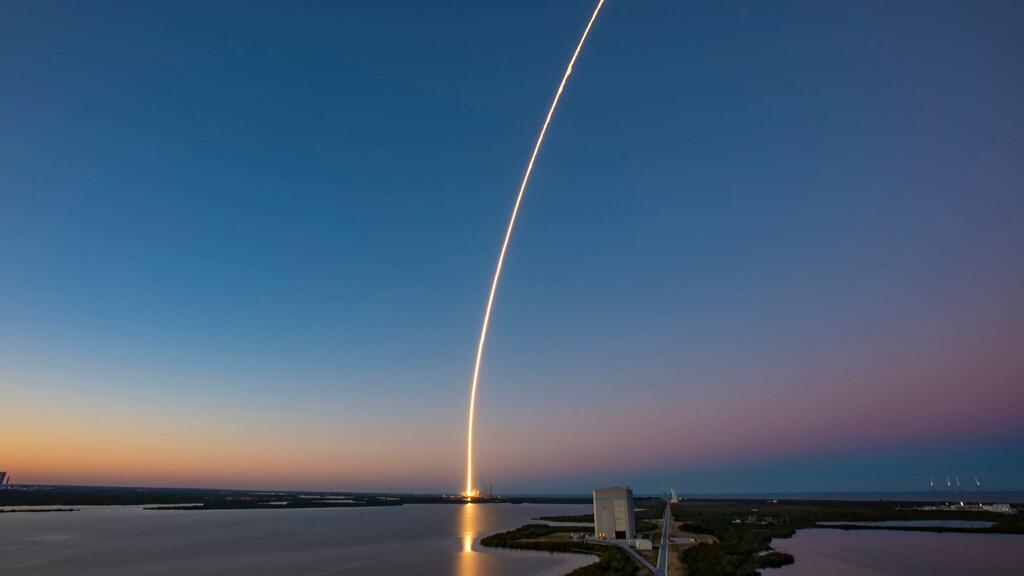 A rocket launches from a coastal launch pad at dusk, leaving a bright curved exhaust trail across the sky above wetlands and water.
