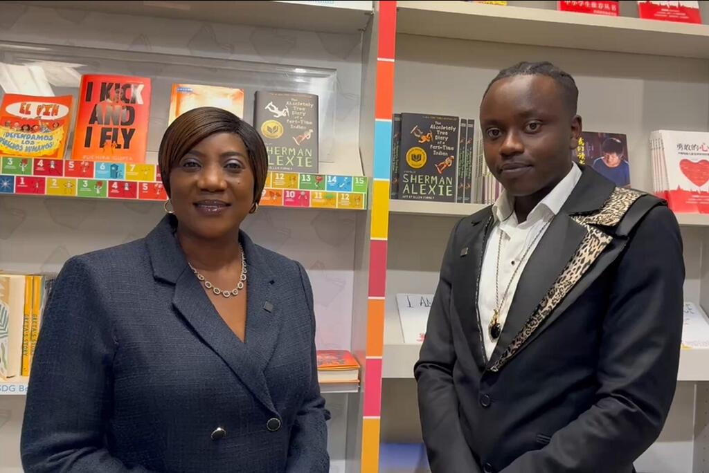 Mwanamke aliyevaa suti ya kijivu na mwanamume aliyevaa koti jeusi lenye lamba la chui wamesimama mbele ya rafu za vitabu zilizojaa vitabu vya Kiingereza na Kichina.