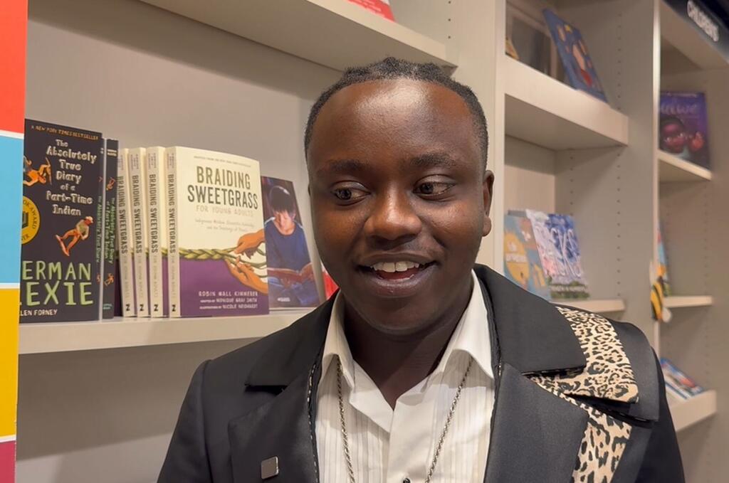 Kijana mweusi aliyevaa koti jeusi na shati jeupe amesimama mbele ya rafu za vitabu akionyesha vitabu vikiwemo Braiding Sweetgrass, akitabasamu kidogo huku akiangalia pembeni.