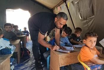 ينحني معلم فوق مكتب خشبي لمساعدة أطفال صغار من الشرق الأوسط على الكتابة في دفاتر داخل فصل دراسي على شكل خيمة مزينة بأعلام وكراسي بلاستيكية.