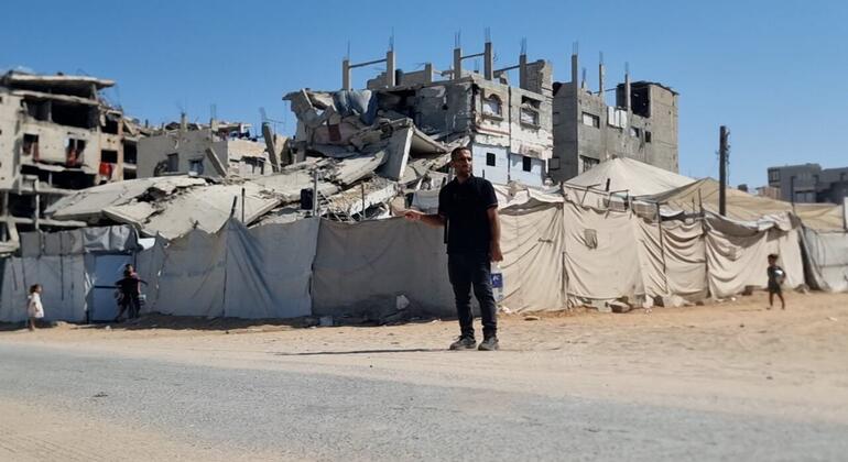 Un homme en chemise noire et pantalon foncé se tient sur une route asphaltée devant des bâtiments fortement endommagés et des rangées de tentes beiges, avec des enfants visibles à proximité sous un ciel bleu clair.