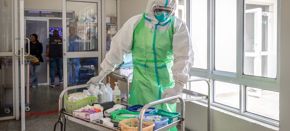 A healthcare worker in white and green personal protective equipment, face shield, mask, and gloves pushes a metal cart stocked with medical supplies and disinfectant bottles through a hospital corridor; people stand near a glass door in the background.