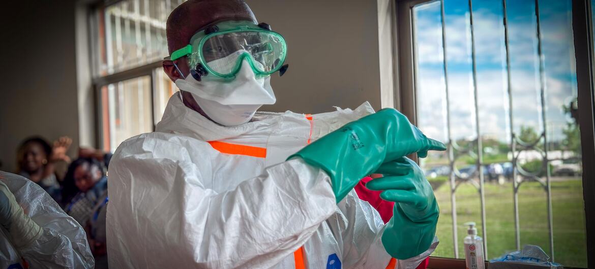 A Black male Kenyan health worker in a white protective suit, face mask, goggles, and green gloves adjusts his glove during a Bundibugyo virus disease response activity.