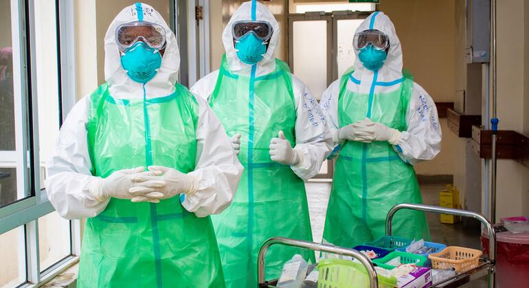 Trois soignants, entièrement équipés de protections individuelles, se tiennent dans un couloir d'hôpital à côté d'un chariot de matériel médical.