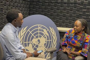 Mwanamume Mweusi aliyevaa shati la bluu nyepesi na mwanamke Mweusi aliyevaa fulana yenye rangi ya juu wakizungumza mbele ya jopo la nembo UMOJA WA MATAIFA wakati wa mahojiano Habari za UN UNGA81.