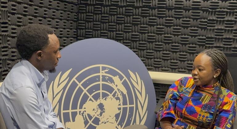 Mwanamume Mweusi aliyevaa shati la bluu nyepesi na mwanamke Mweusi aliyevaa fulana yenye rangi ya juu wakizungumza mbele ya jopo la nembo UMOJA WA MATAIFA wakati wa mahojiano Habari za UN UNGA81.