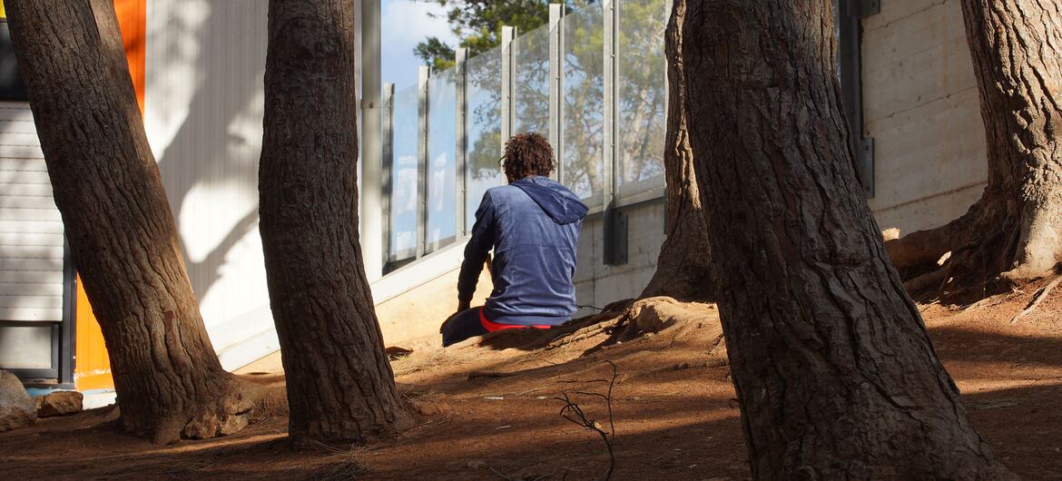 A young unaccompanied child sits alone on dirt among tree trunks outside a fenced building at a migrant reception centre in Lampedusa.