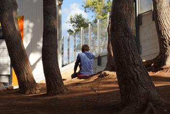 A young unaccompanied child sits alone on dirt among tree trunks outside a fenced building at a migrant reception centre in Lampedusa.