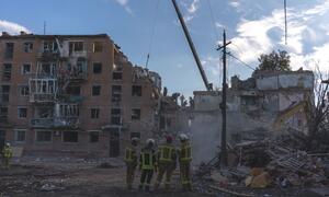Rescue workers in yellow reflective uniforms and helmets inspect rubble outside a heavily damaged multi-story brick residential building; a crane arm and excavator are visible amid debris.