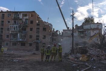 Des secouristes en uniformes et casques jaunes réfléchissants inspectent les décombres à l'extérieur d'un immeuble résidentiel en briques de plusieurs étages fortement endommagé ; un bras de grue et une excavatrice sont visibles au milieu des débris.