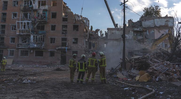 Des secouristes en uniformes et casques jaunes réfléchissants inspectent les décombres à l'extérieur d'un immeuble résidentiel en briques de plusieurs étages fortement endommagé ; un bras de grue et une excavatrice sont visibles au milieu des débris.