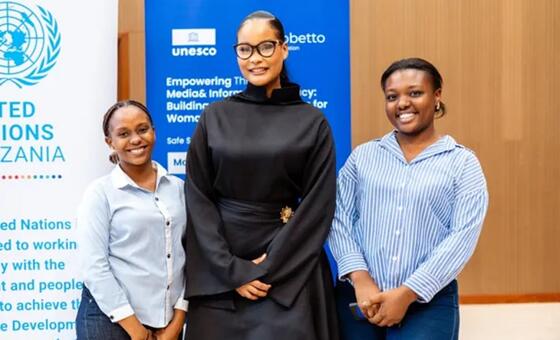 Wanawake watatu wenye tabasamu wamesimama mbele ya bango la bluu UNESCO na UMOJA WA MATAIFA Tanzania lenye maandishi kuhusu kuwawezesha wanawake kupitia vyombo vya habari na usalama wa habari.