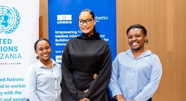 Wanawake watatu wenye tabasamu wamesimama mbele ya bango la bluu UNESCO na UMOJA WA MATAIFA Tanzania lenye maandishi kuhusu kuwawezesha wanawake kupitia vyombo vya habari na usalama wa habari.