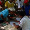 Des hommes et des femmes noirs se rassemblent autour d'une table à l'extérieur tandis que des travailleurs en chemises blanches ornées d'écussons du drapeau haïtien signent et vérifient les formulaires de distribution de semences.