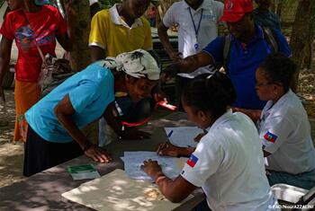 Des hommes et des femmes noirs se rassemblent autour d'une table à l'extérieur tandis que des travailleurs en chemises blanches ornées d'écussons du drapeau haïtien signent et vérifient les formulaires de distribution de semences.