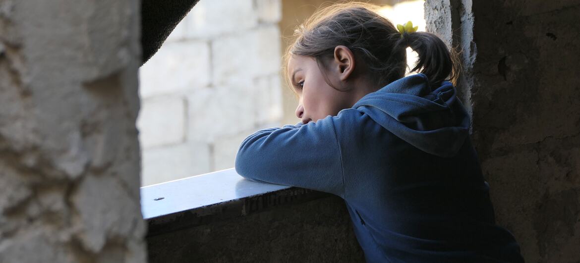 A young Middle Eastern girl in a blue hooded sweatshirt rests her arms on a concrete windowsill and gazes outside from a dim room in Aleppo.