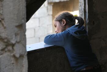 À Alep, une jeune fille du Moyen-Orient, vêtue d'un sweat-shirt à capuche bleu, pose ses bras sur le rebord d'une fenêtre en béton et regarde dehors depuis une pièce faiblement éclairée.