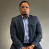 Mwanamume Mweusi aliyevaa suti ya kijivu na shati la bluu nyepesi ameketi kwenye mandhari ya studio ya kijivu isiyo na rangi, akiwa amevaa miwani isiyo na rimu na maikrofoni ndogo ya lavalier.