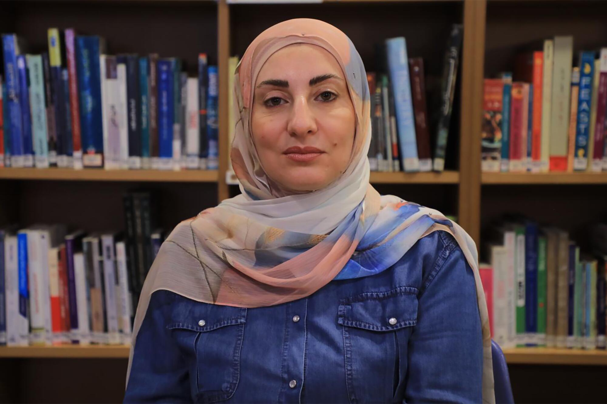وفاء شرارة، عضوة في لجنة أمينات المكتبات النسائية في مكتبة بنت جبيل، ترتدي الحجاب والقميص الأزرق أمام رفوف الكتب في المكتبة.